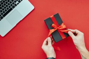 Hands tying a red ribbon on a gift box beside a laptop on a red surface.