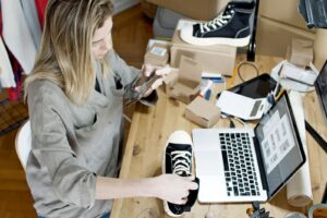 Small business owner photographing a black sneaker beside a laptop and shipping boxes.