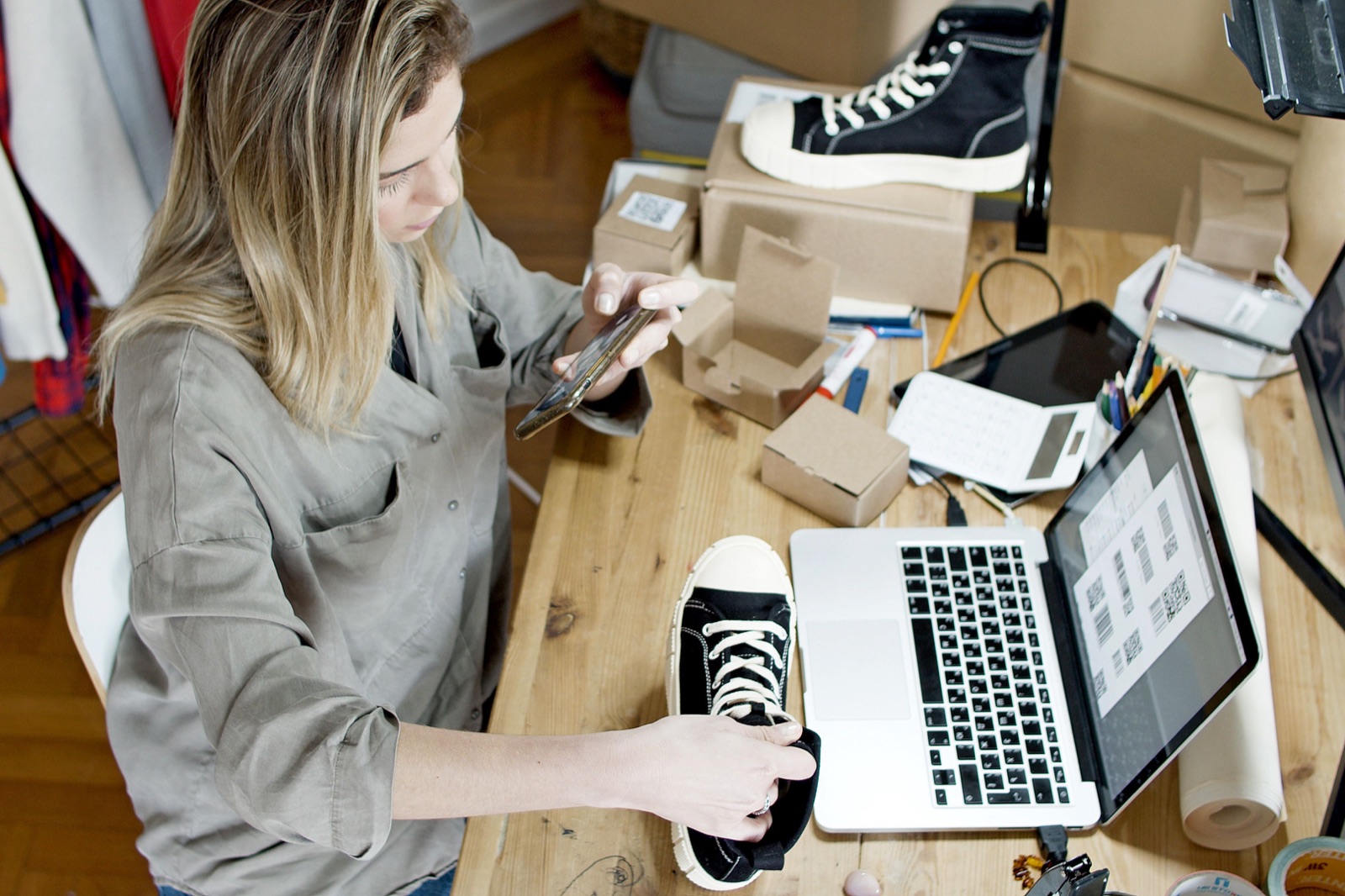 Small business owner photographing a black sneaker beside a laptop and shipping boxes.