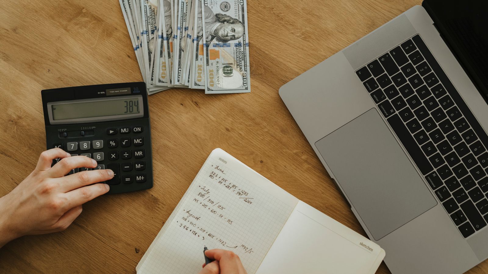 Hands using a calculator beside a laptop and finance notebook.